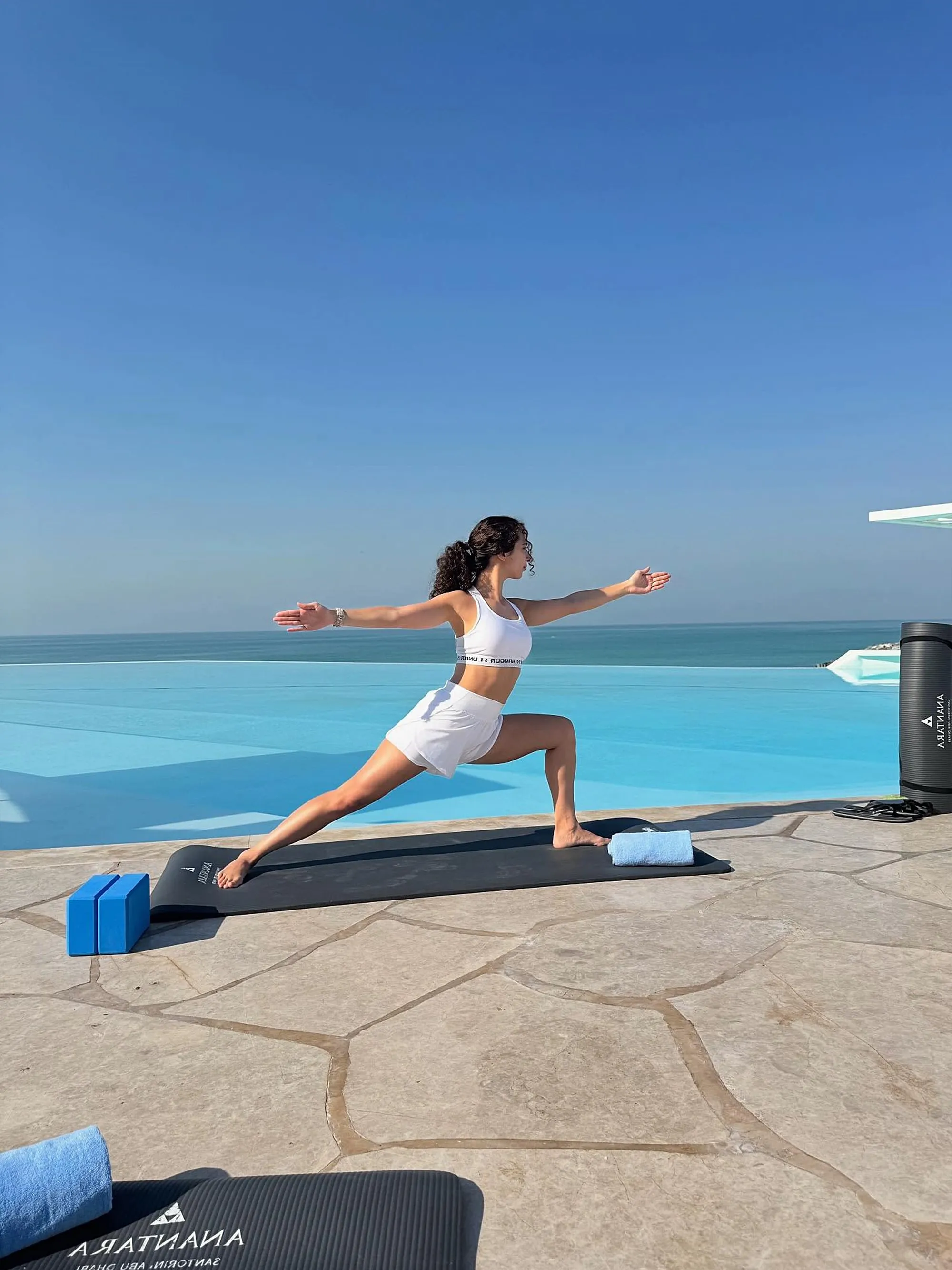 Teresa Villa holding a standing lunge with her arms stretched wide, on a terrace beside an infinity pool and the sea